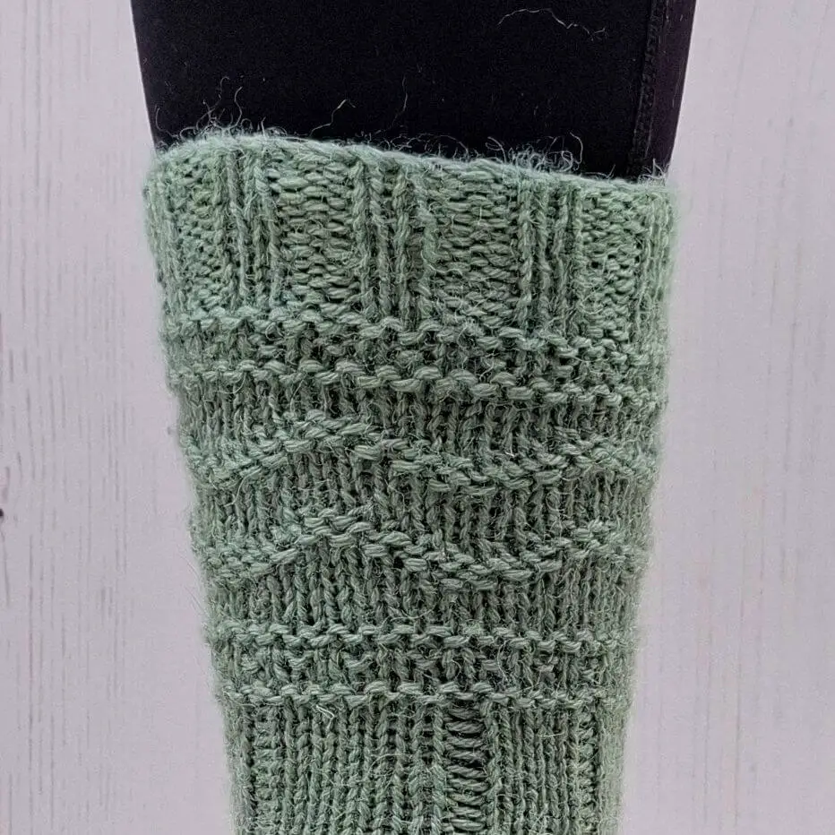 A close-up of the pattern of a green knitted sock on a leg. The knit features ribbing and textured patterns, set against a light background.