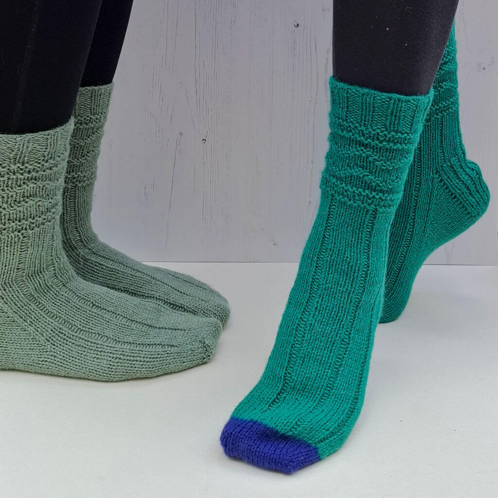People wearing knitted socks: one pair is light green, the other bright green with a blue toe. The background is a white wooden texture.