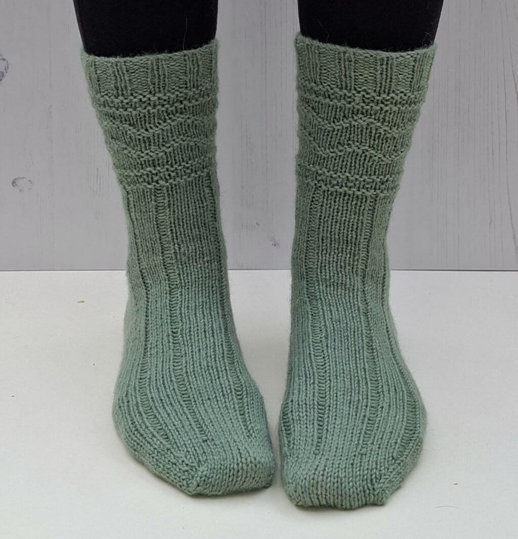 Green knitted socks on feet against a light wooden background. The socks feature a ribbed and textured pattern.