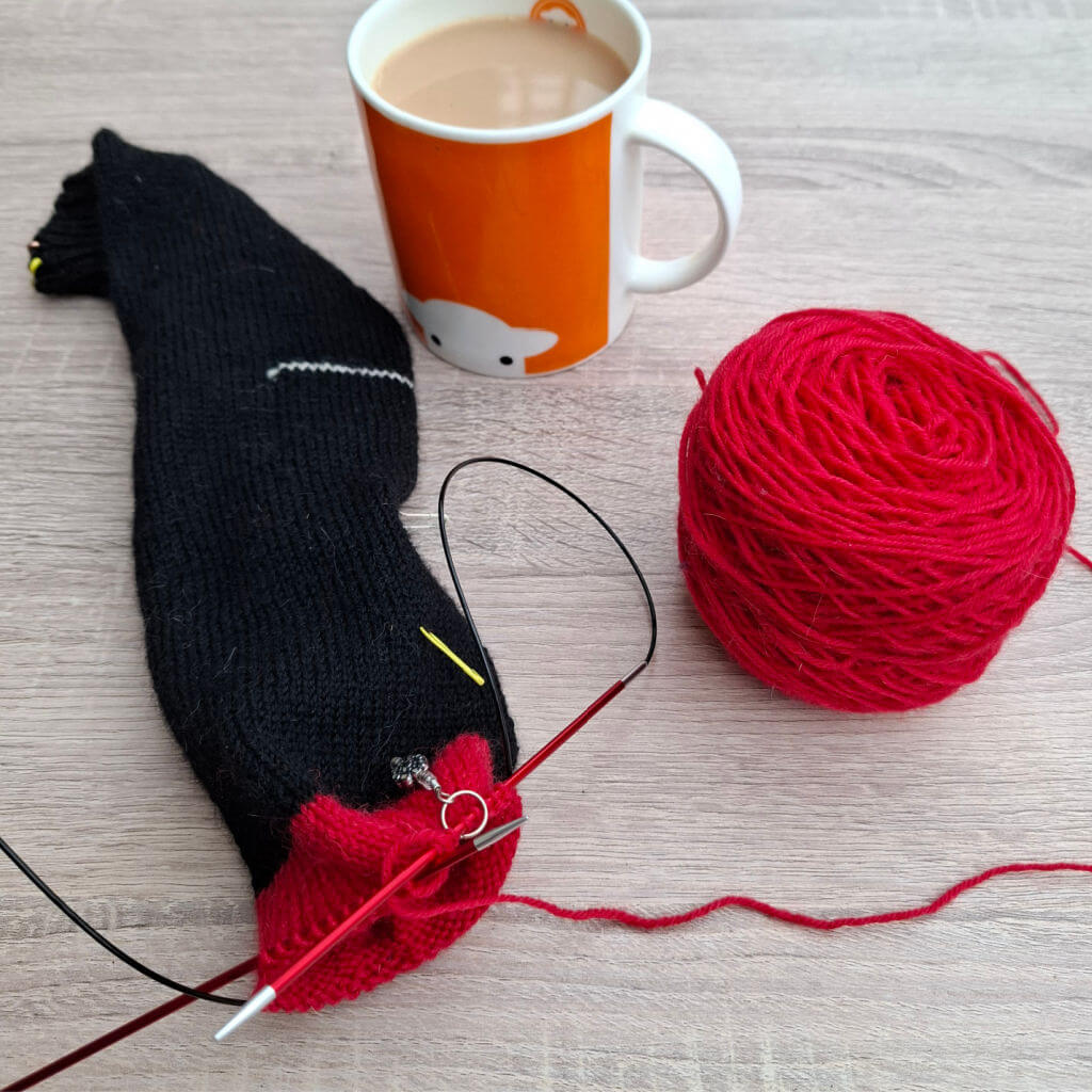Knitting scene with a partially knitted black sock, red yarn ball, and circular knitting needles on a wooden table. An orange mug of tea is beside them.