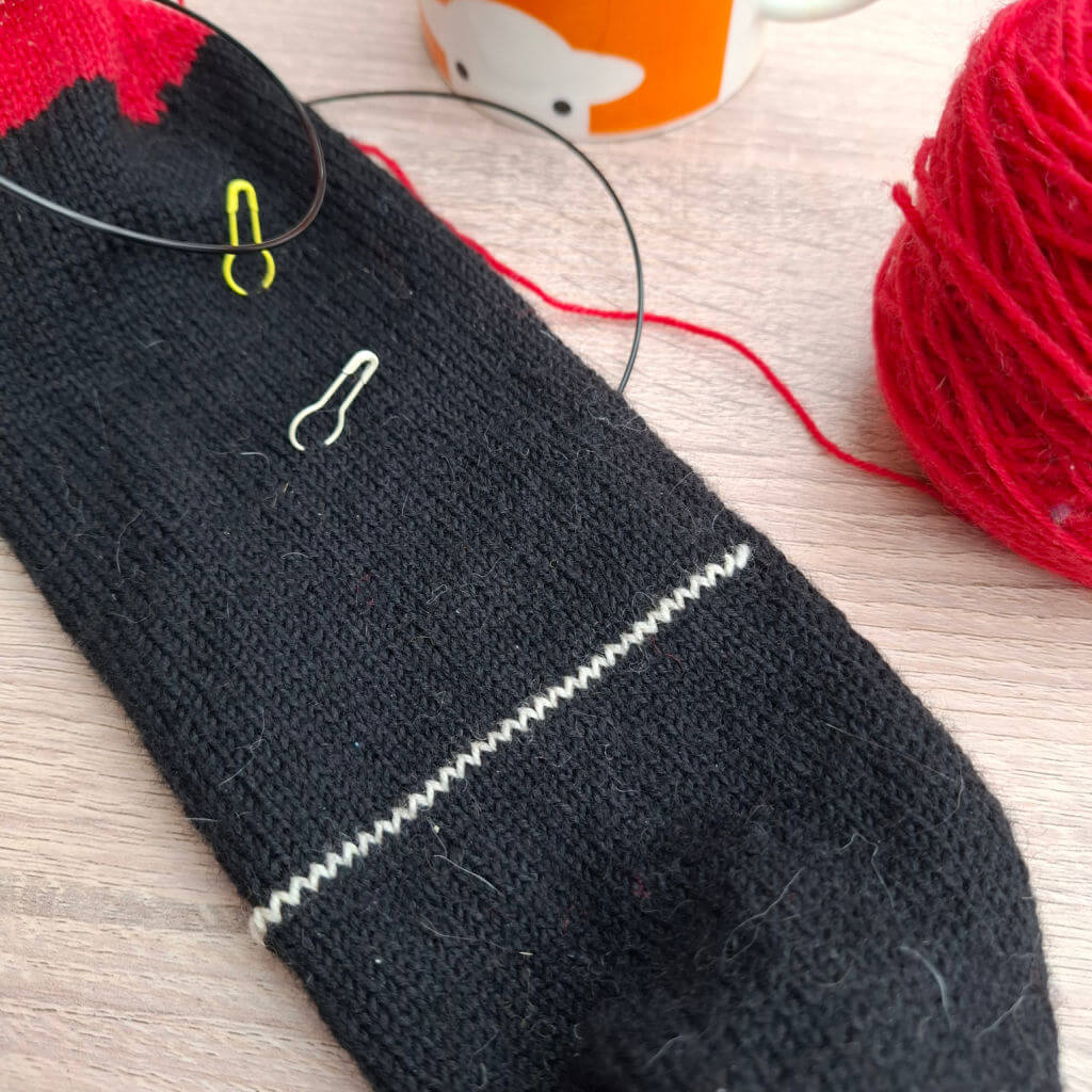 Knitted black sock with red heel and contrasting white stitching laid on a wooden surface. Two stitch markers are attached. Red yarn ball and mug nearby.