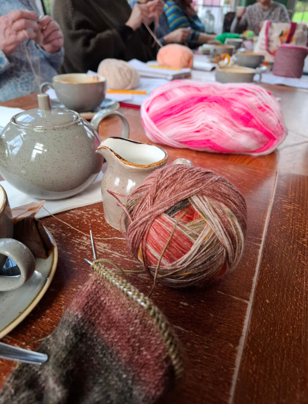 A cosy knitting circle on a wooden table with colourful yarn, knitting needles, teapot, and cups. Participants focus on their craft, creating a warm, creative atmosphere.