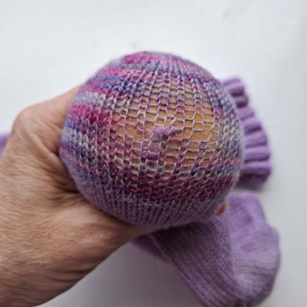 A hand holds a purple and pink knitted sock in the process of being darned, with visible mesh stitches and a wooden darning tool inside.