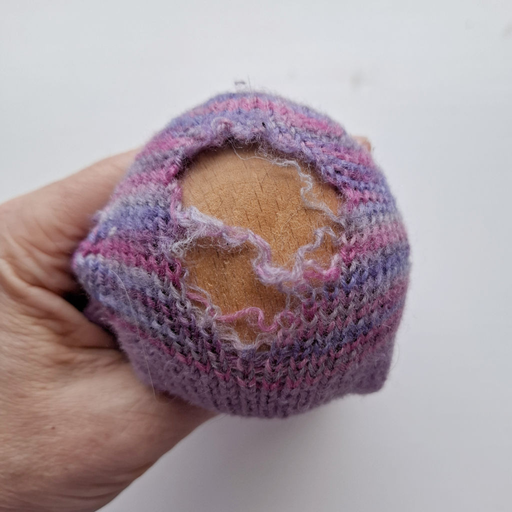 A hand holds a wooden darning egg covered by a purple and pink sock with a large hole, showing frayed edges. The tone suggests repair work.