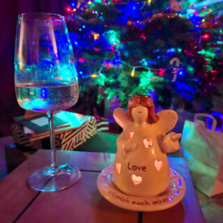 A wine glass and an angel figurine labeled "Love" sit on a table, set against a backdrop of a brightly lit Christmas tree, creating a festive atmosphere.