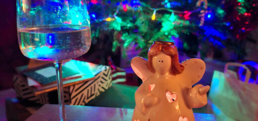 A wine glass and an angel figurine labeled "Love" sit on a table, set against a backdrop of a brightly lit Christmas tree, creating a festive atmosphere.