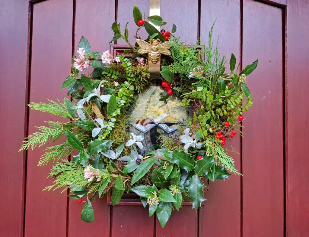 Festive wreath on a red door, featuring green foliage, red berries, pink flowers, and a gold bee ornament. It conveys a cheerful, holiday atmosphere.
