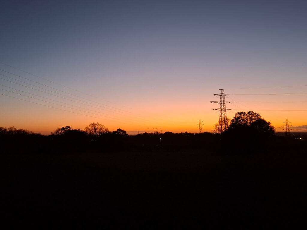 Sunset with a gradient sky, orange fading to deep blue. Silhouetted trees and power lines create a serene, tranquil scene.