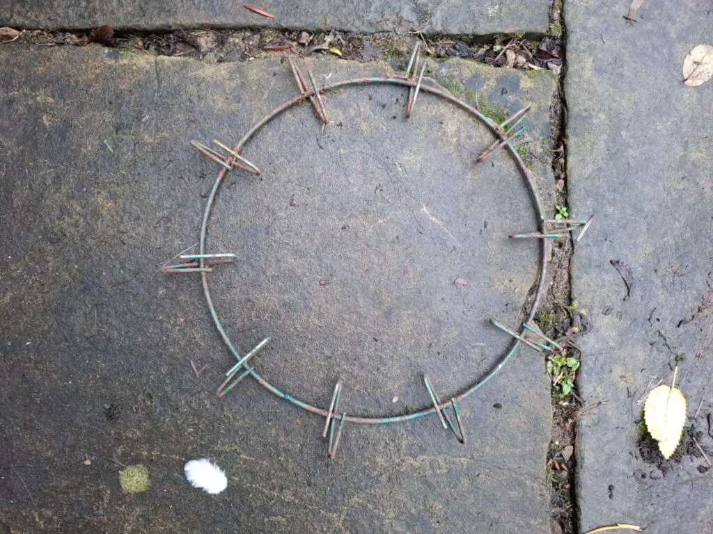 A rusty metal ring with evenly spaced clips lies on a weathered stone pavement, surrounded by moss and a few scattered leaves, conveying an aged, earthy feel.