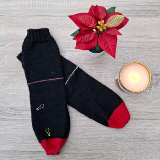 Black socks with red toes and small colourful paperclips as row markers lie on a wooden surface. Beside them, a lit candle and a red poinsettia add warmth and festivity.