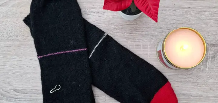 Black socks with red toes and small colourful paperclips as row markers lie on a wooden surface. Beside them, a lit candle and a red poinsettia add warmth and festivity.