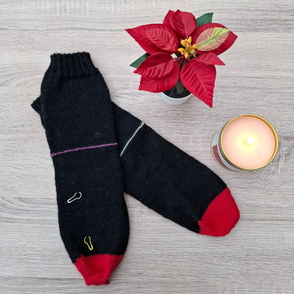 Black socks with red toes and small colourful paperclips as row markers lie on a wooden surface. Beside them, a lit candle and a red poinsettia add warmth and festivity.