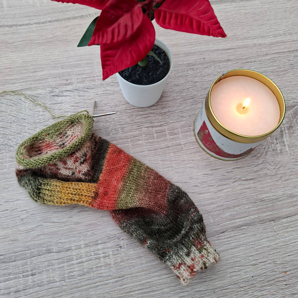 Colourful knitted sock with autumn tones lies on a wooden table next to a lit candle in a tin and a small potted red poinsettia plant. Cosy atmosphere.