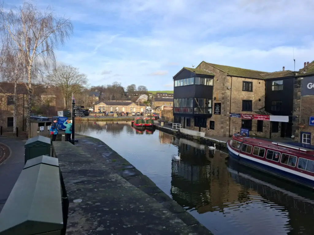 Canal scene with stone buildings, moored boats, and a tree under a blue sky. A bridge in the foreground and distant hills add depth and tranquility.