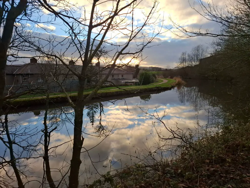 A serene canal reflects the cloudy sky at sunset, surrounded by bare trees. Houses line the water, and a path curves alongside, creating a peaceful rural scene.