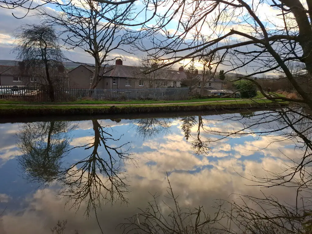 A serene canal reflects bare trees and a cloudy sky at sunset. Houses line the opposite bank, framed by branches, creating a peaceful, picturesque scene.