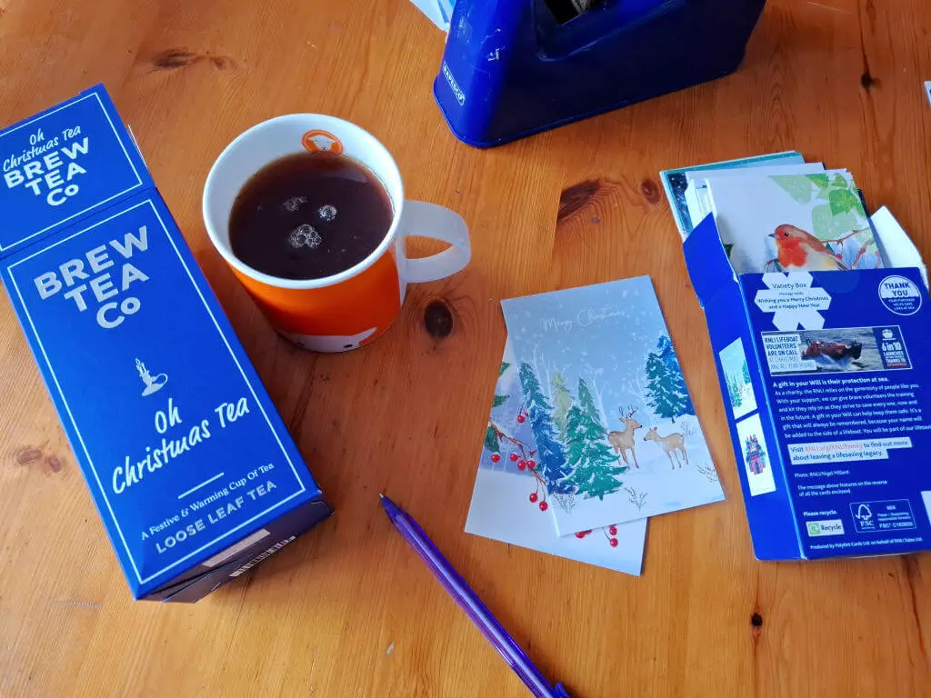 A cosy scene shows a blue tea box, a cup of tea, and Christmas cards on a wooden table. The mood is warm and festive, evoking holiday preparation.
