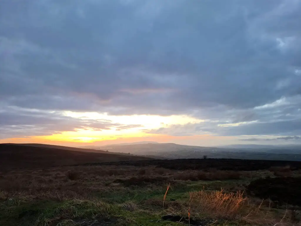 Expansive landscape at sunset with rolling hills and dark clouds. The sun casts a warm glow, creating a serene, peaceful atmosphere.