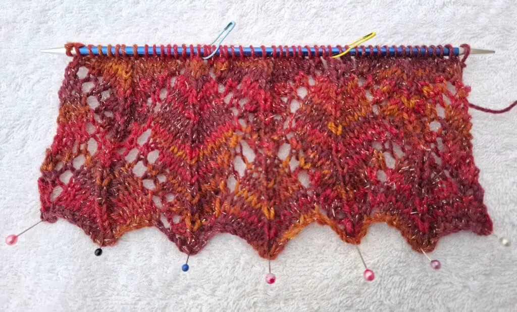 A piece of knitting in progress on a needle features a lace pattern with red, orange, and brown yarn. Pin markers highlight sections on the white background.