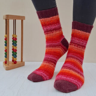 Person wearing vibrant, striped socks in red, orange, and pink hues stands on a light wooden floor. An abacus with colourful beads is next to them.