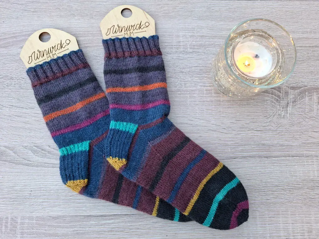 Colourful striped socks on a wooden background beside a lit candle, creating a cosy and warm ambiance. Socks feature blue, pink, and orange hues.