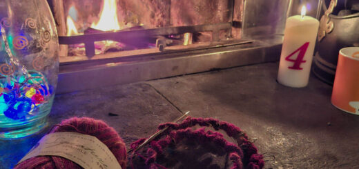Cosy scene with knitting supplies, colourful yarn in front of a warm fireplace. A lit candle marked "4" and vibrant glass decor set a relaxing winter mood.