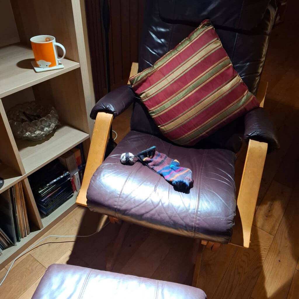A cosy room features a leather chair with a striped pillow and a partly-knitted sock on it. A wooden shelf holds a sheep mug and books. Warm, inviting atmosphere.