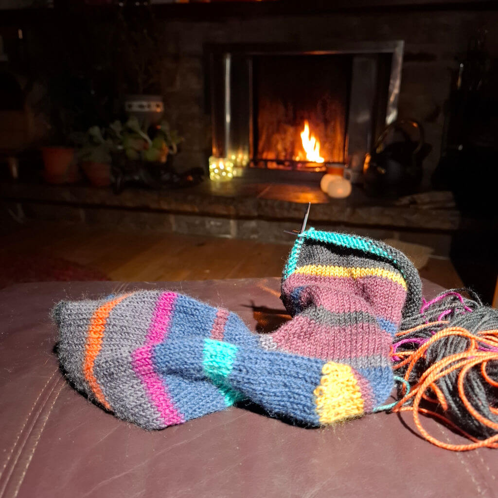 Colourful knitted socks and yarn on a leather footstool, with a cosy lit fireplace in the background, creating a warm, inviting atmosphere.