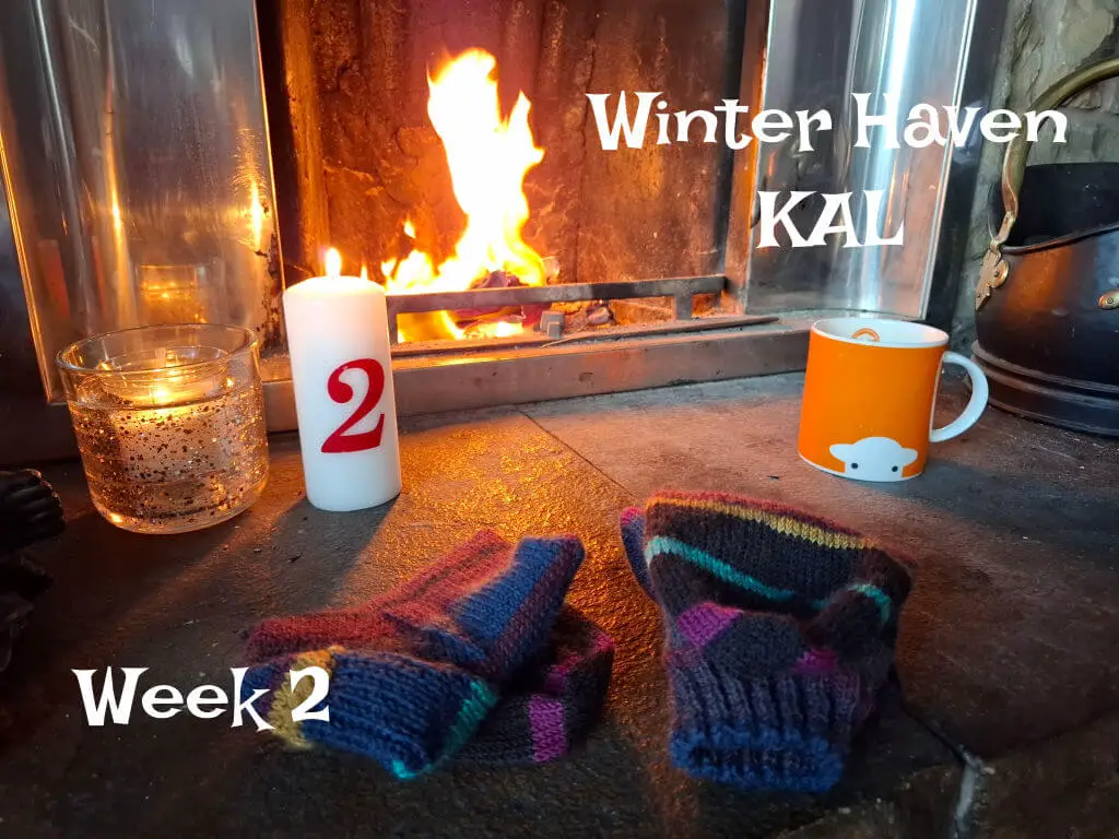 Cosy scene by a lit fireplace with striped knit socks, a red-numbered candle, and an orange mug. Text reads, "Winter Haven KAL, Week 2."