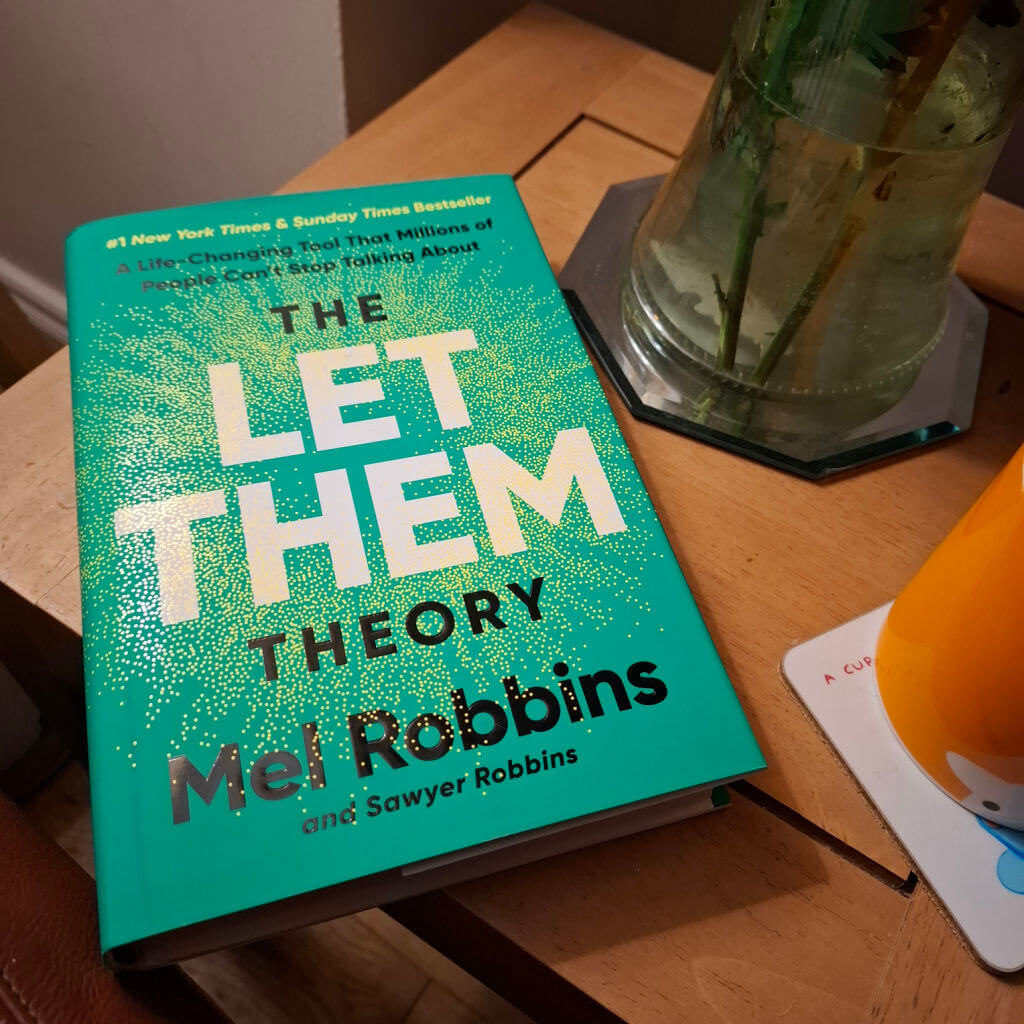 A teal book titled "The Let Them Theory" by Mel Robbins rests on a wooden table, near a vase with water and flower stalks visible. The scene feels calm and inviting.