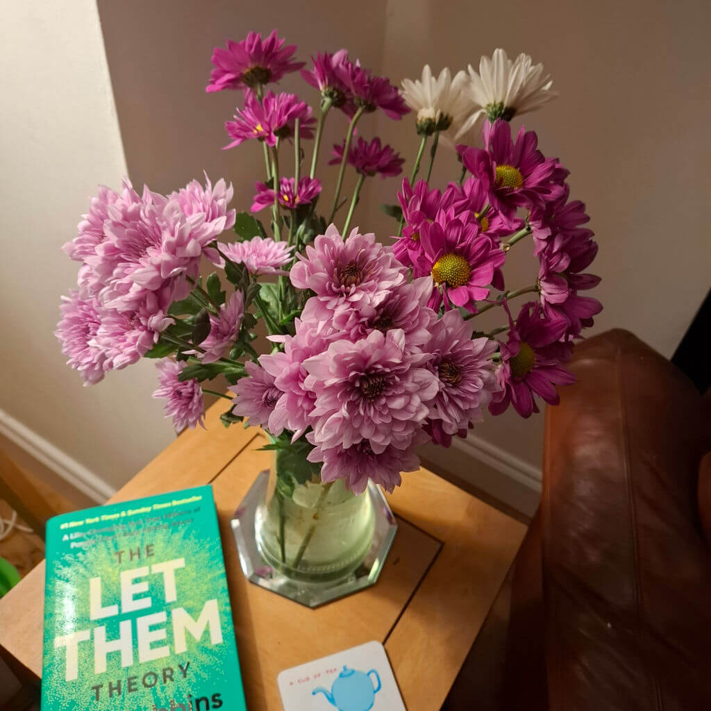 A vase with pink and white flowers sits on a wooden table beside a teal book titled "The Let Them Theory" and a coaster with a teapot design. Cosy ambiance.
