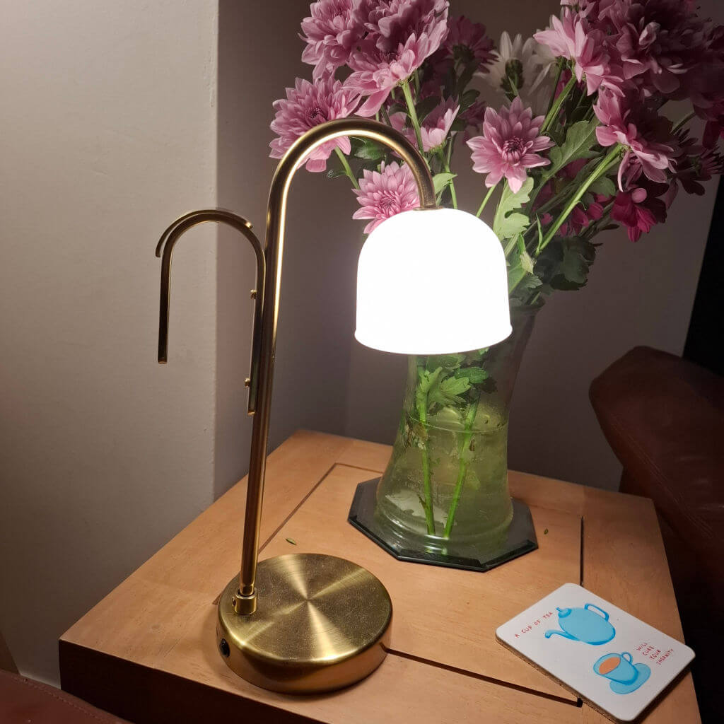 A cosy room corner with a brass table lamp casting a warm glow. Next to it, a vase of pink flowers and a coaster featuring a blue teapot and cup.