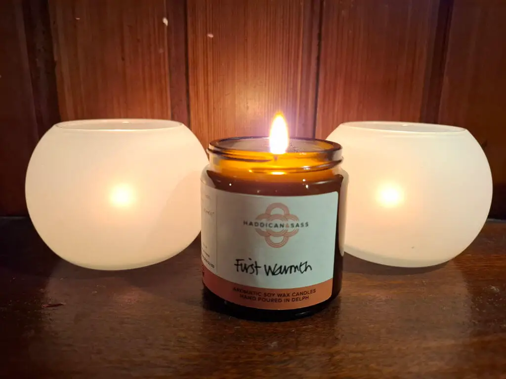 A lit candle in an amber glass jar labeled "First Warmth" is flanked by two glowing white candle holders on a wooden surface, creating a cosy ambiance.