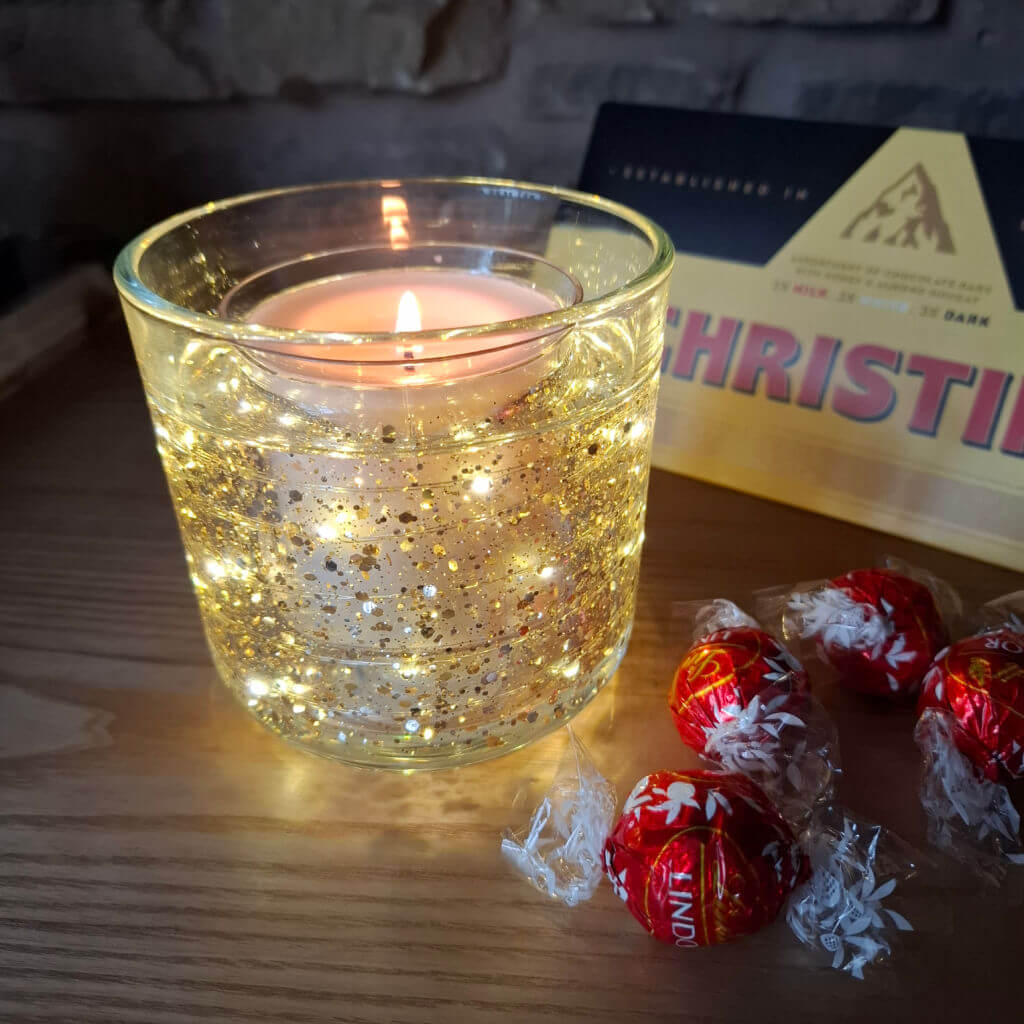 A lit candle in a glass with golden glitter, creating a warm glow. Nearby are wrapped red chocolates and a chocolate box on a wooden table.