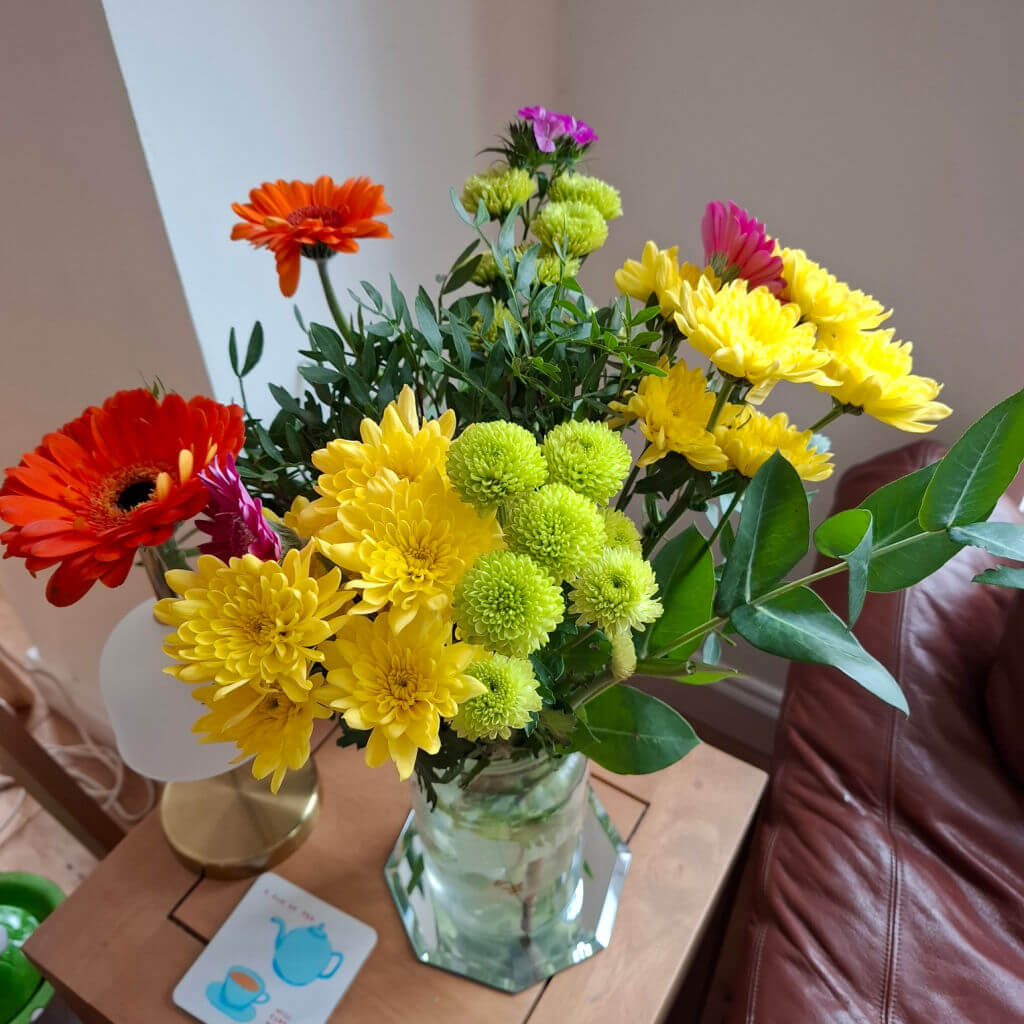 A vibrant bouquet of yellow, orange, red, and pink flowers in a glass vase on a wooden table, creating a cheerful and lively atmosphere.