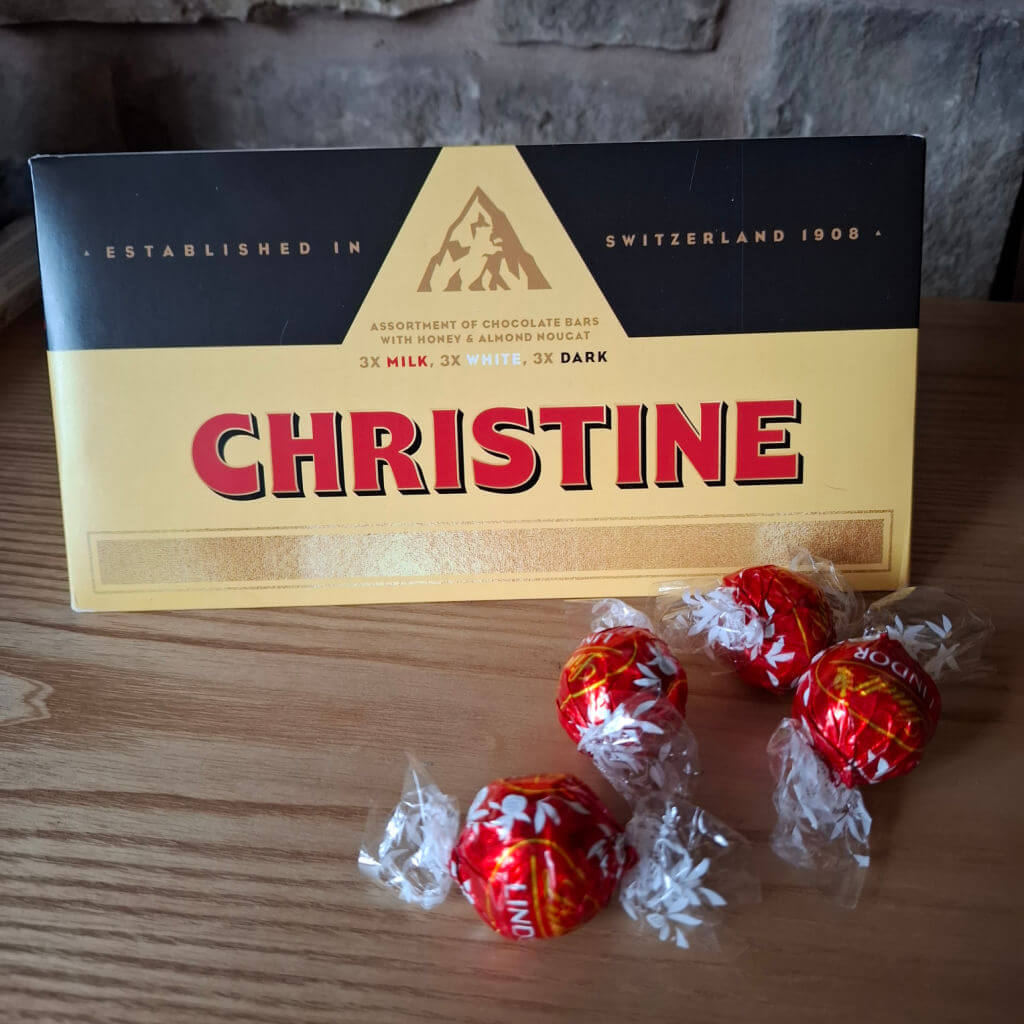 A box of Swiss Toblerone chocolate with "CHRISTINE" in bold red letters sits on a wooden table, surrounded by four wrapped red and white Lindor truffles.