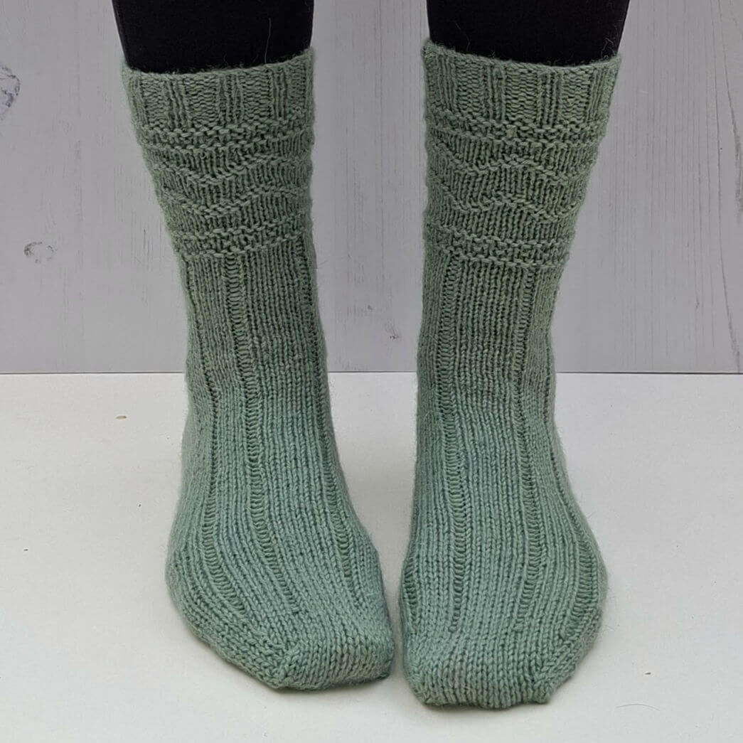 Green knitted socks on feet against a light wooden background. The socks feature a ribbed and textured pattern.