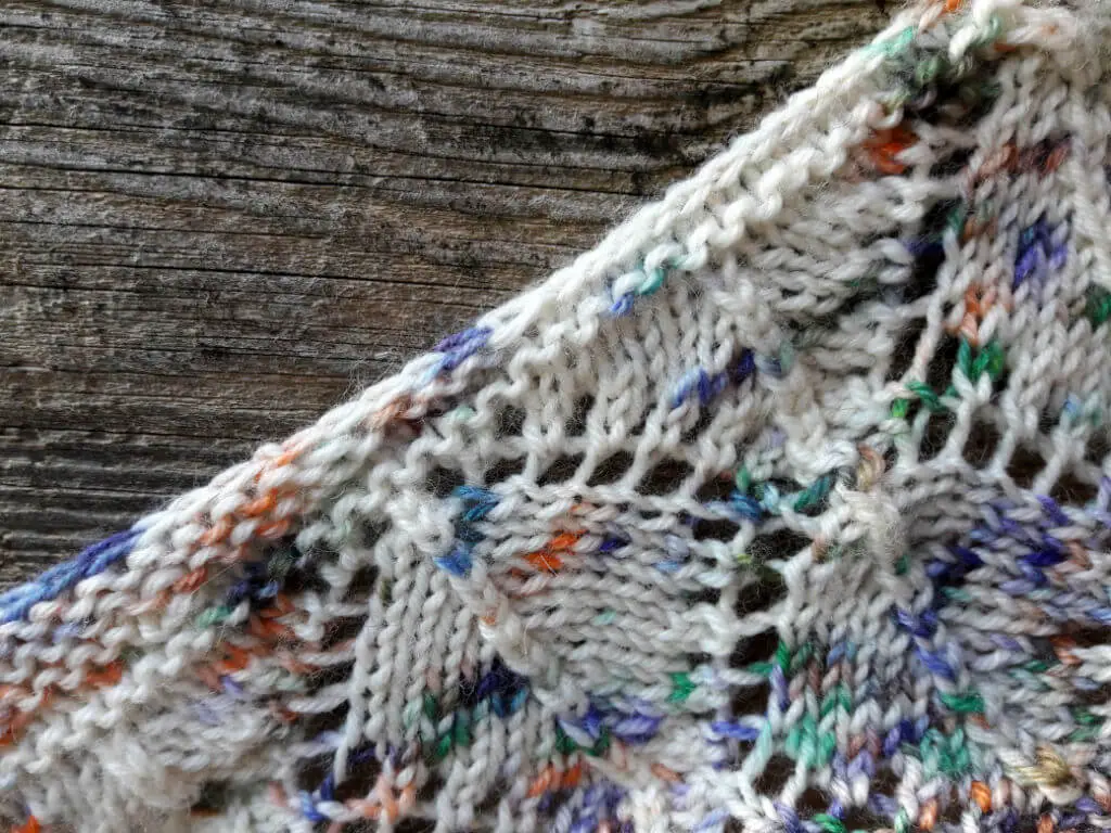 Close-up of a knitted fabric with multicoloured yarn, featuring white, orange, green, and blue tones, against a rustic wooden fence.