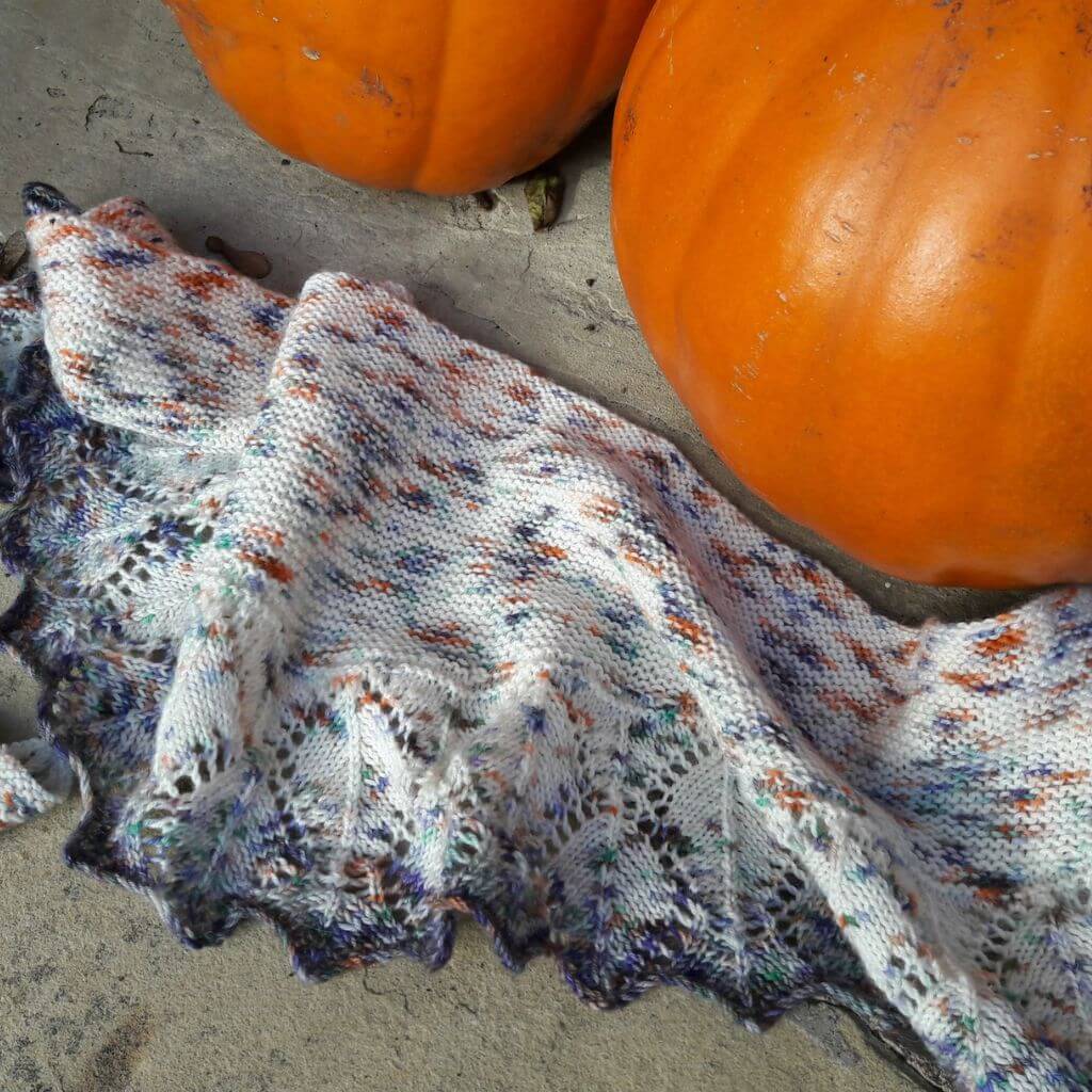 A colourful knitted shawl with autumnal hues of orange and blue is draped on stone flags next to two large, round pumpkins, evoking a cosy autumn scene.