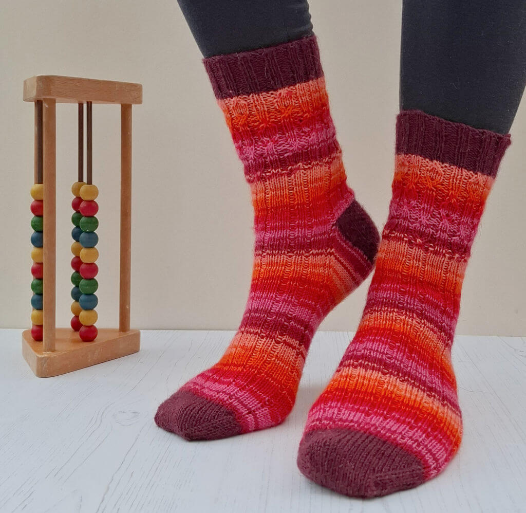 Person wearing vibrant, striped socks in red, orange, and pink hues stands on a light wooden floor. An abacus with colourful beads is next to them.
