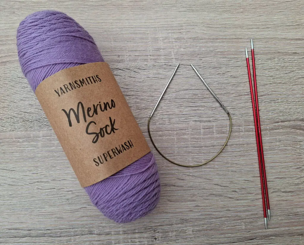 A skein of purple "Merino Sock" yarn lies on a wooden surface, flanked by red knitting needles and circular metal needles, conveying a cosy, crafting vibe.