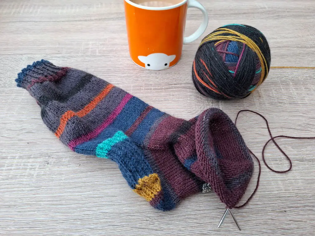Knitted multicoloured sock lies on a wooden table, with a ball of yarn and knitting needles beside it. An orange mug with a sheep design sits nearby. Cosy atmosphere.