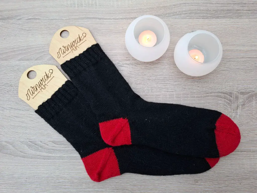 Black socks with red toes and heels are displayed on wooden sock blockers on a light wooden surface. Two lit candles in white holders add a cosy ambiance.
