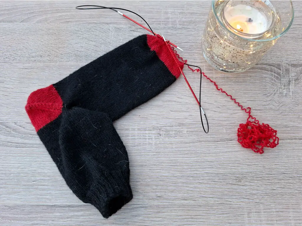 A partially knitted black sock with red heel and toes is on wooden table. Knitting needles and yarn are attached. A glowing, decorated candle is beside it.