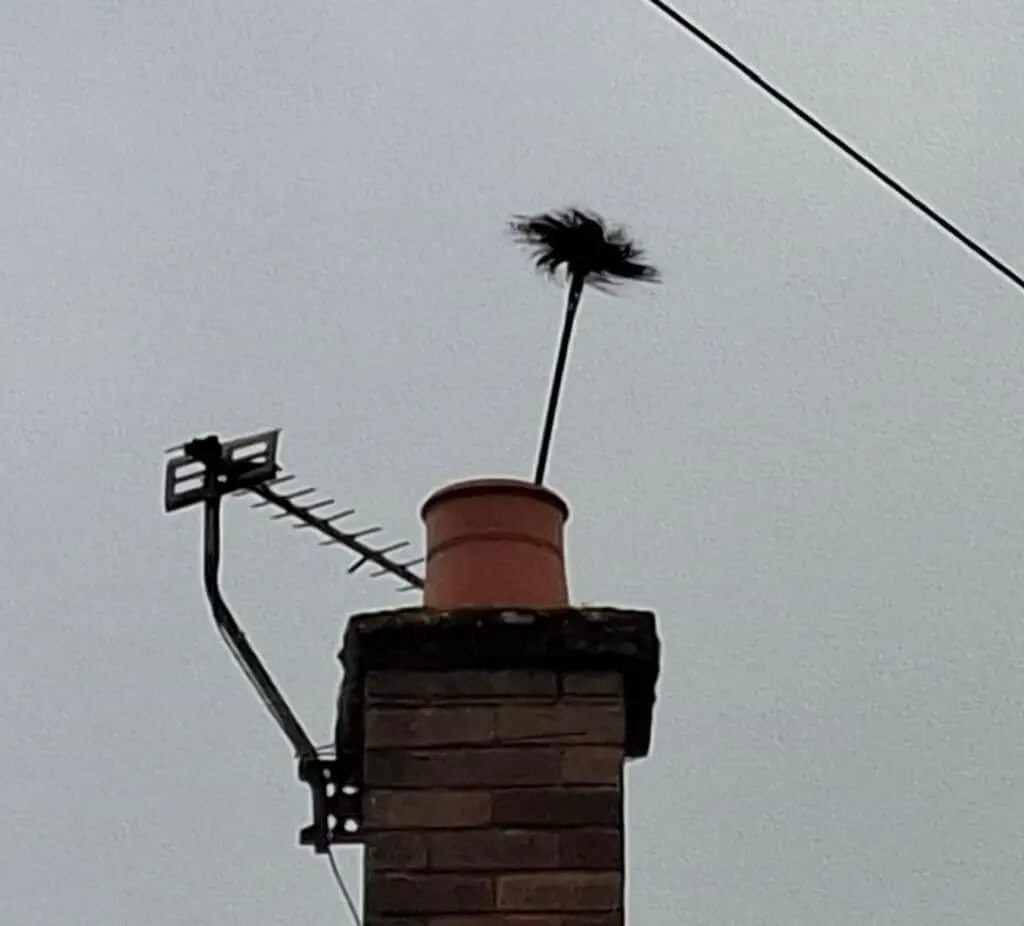 A black chimney sweep's brush emerging from a chimney pot, next to a TV antenna, under an overcast sky.