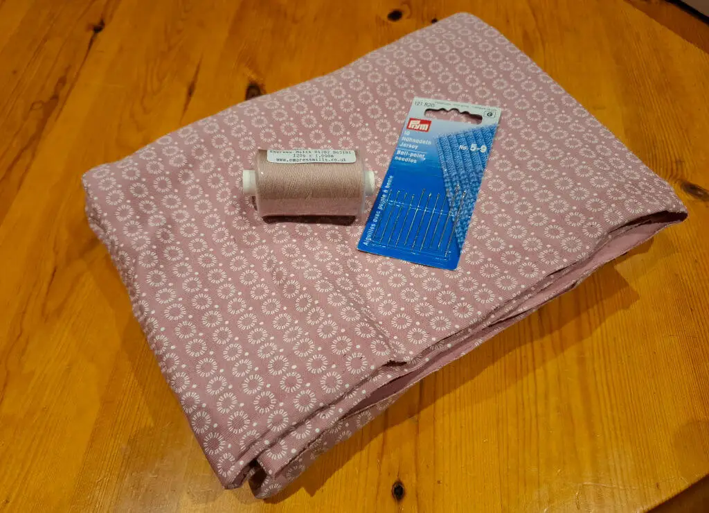 Pink fabric with a white floral pattern is neatly folded on a wooden table. A brown thread spool and a packet of sewing needles are placed on top.
