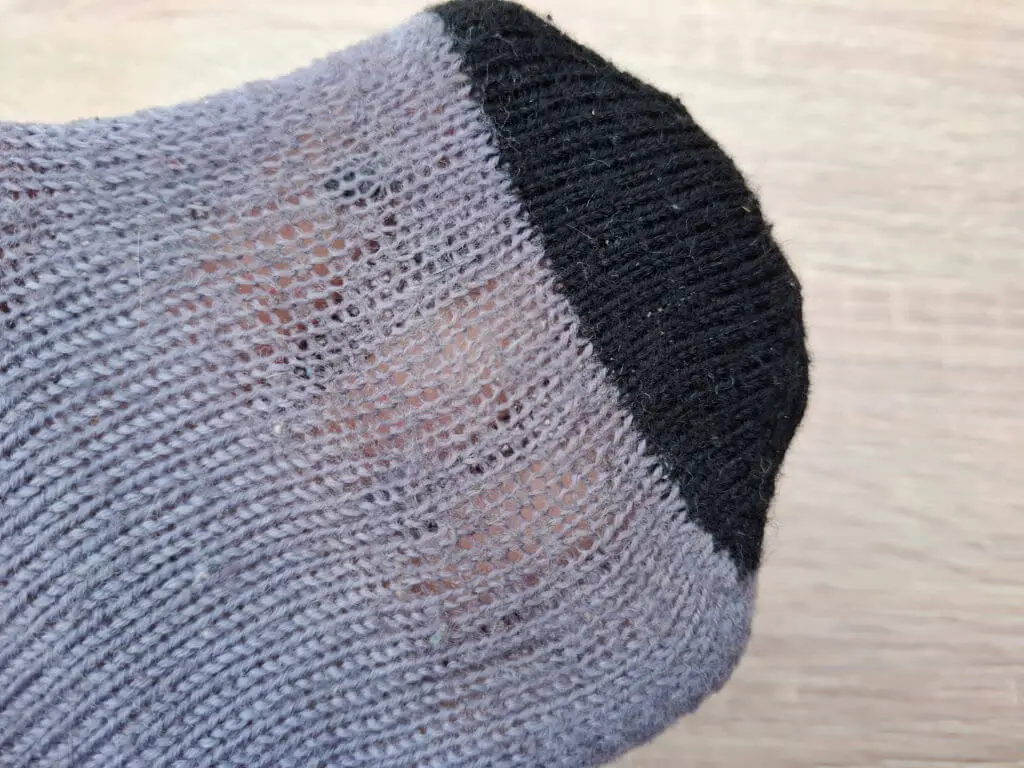 Close-up of a worn purple sock with a black toe area. The fabric is thinning, revealing skin beneath, indicating heavy use. Background is a light wood floor.