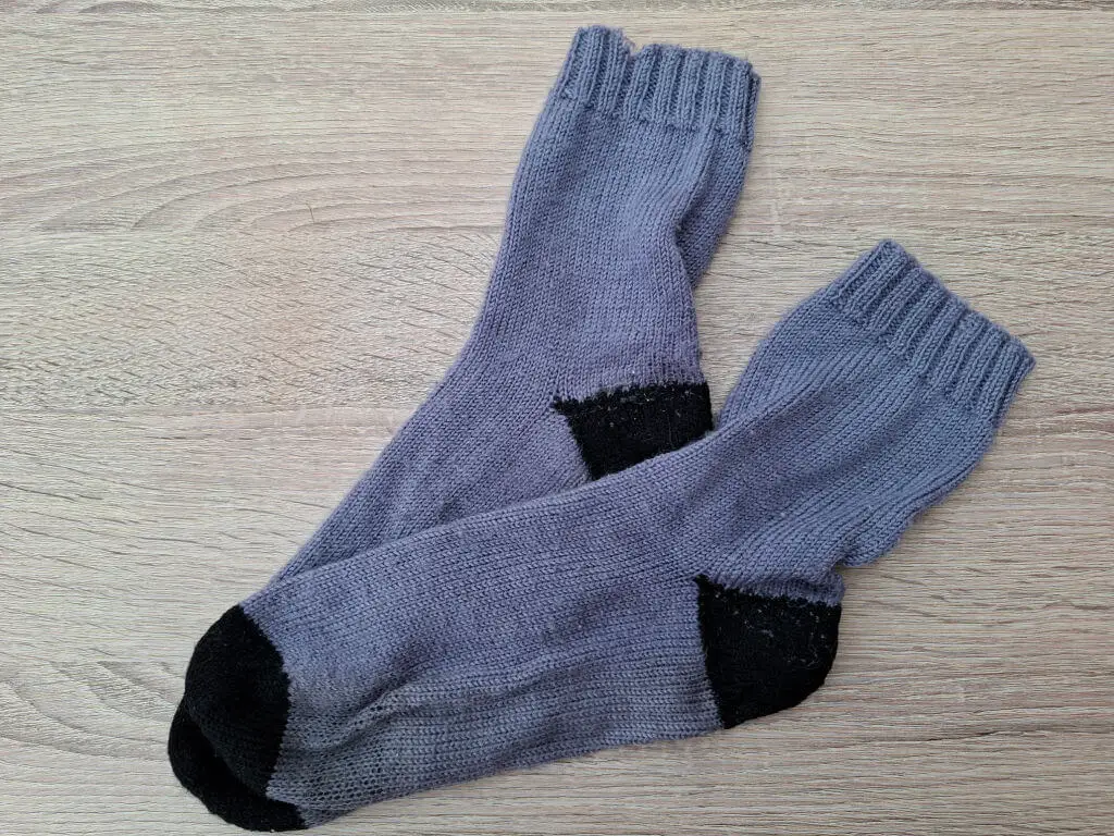 A pair of purple knitted socks with black heels and toes lies on a light wooden surface. The socks are slightly overlapping, creating a cosy appearance.