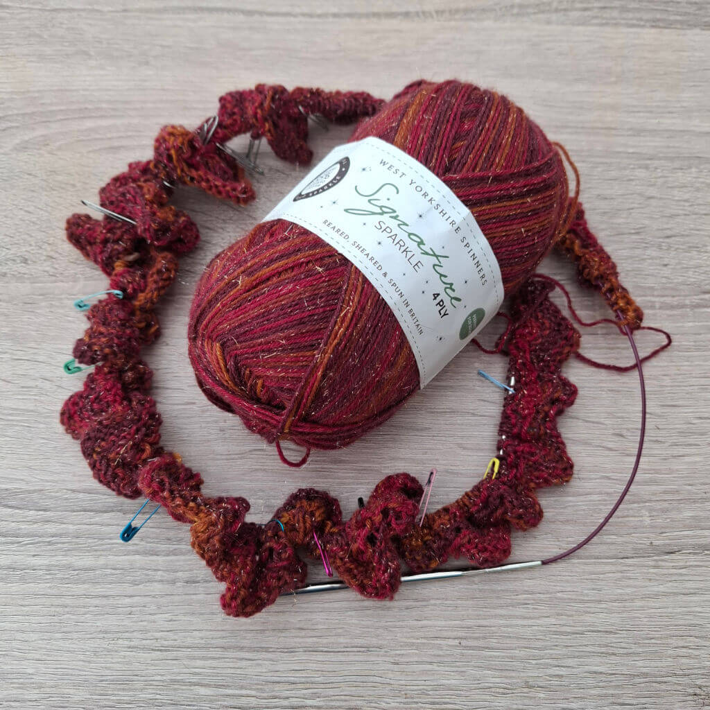 A ball of red and gold sparkly yarn labeled "Signature Sparkle 4 Ply" next to a partially knitted shawl with colourful stitch markers on a wooden table.