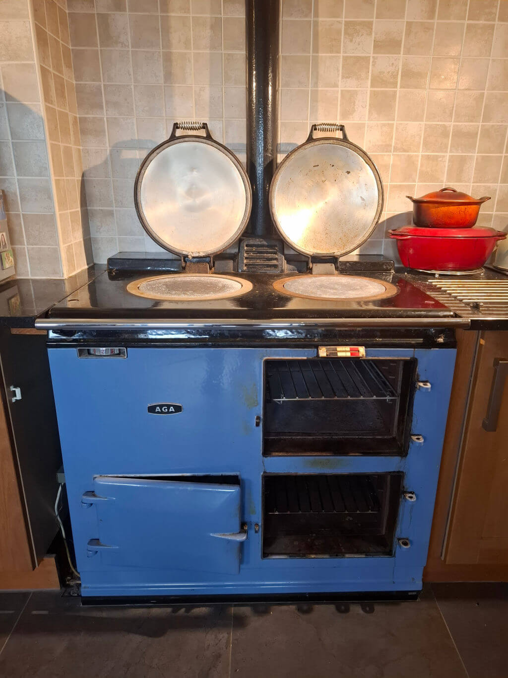Blue AGA stove with dual hotplates open, silver lids reflecting light. Red pot on right. Tile backsplash and wooden cabinets frame the scene. Cosy and vintage.